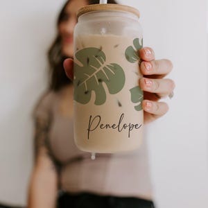 K&ouml;nnte beinhalten: Ein durchsichtiger Glasbecher mit Holzdeckel und Strohhalm, gef&uuml;llt mit einem hellbraunen Getr&auml;nk. Der Becher hat ein gr&uuml;nes Monstera-Blatt-Design und den Namen "Penelope" in Schreibschrift. Das Getr&auml;nk scheint Eiskaffee zu sein.