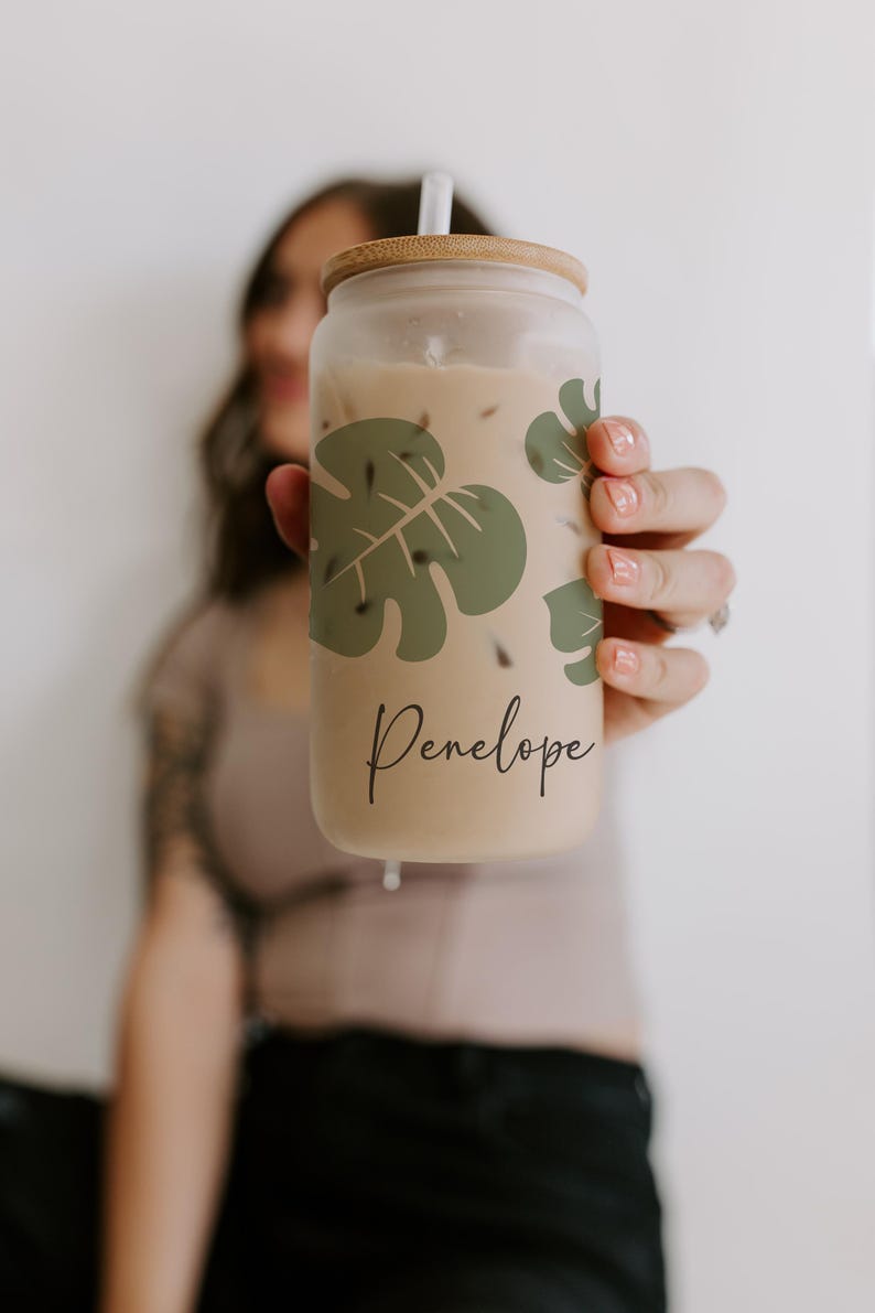 K&ouml;nnte beinhalten: Ein durchsichtiger Glasbecher mit Holzdeckel und Strohhalm, gef&uuml;llt mit einem hellbraunen Getr&auml;nk. Der Becher hat ein gr&uuml;nes Monstera-Blatt-Design und den Namen "Penelope" in Schreibschrift. Das Getr&auml;nk scheint Eiskaffee zu sein.