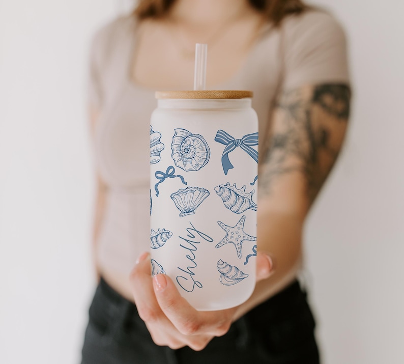 K&ouml;nnte beinhalten: Ein Glasbecher mit Bambusdeckel und Strohhalm, verziert mit blauen Muschel- und Schleifenillustrationen. Der Name "Shelly" ist in einer Schreibschrift geschrieben. Der Becher wird vor einer Person gehalten.