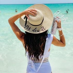 May include: A woman wearing a white swimsuit with a sash that says "Bride" and a straw hat that says "Mrs. DeWitt". She is holding a white tumbler with a straw that says "Mrs. DeWitt".