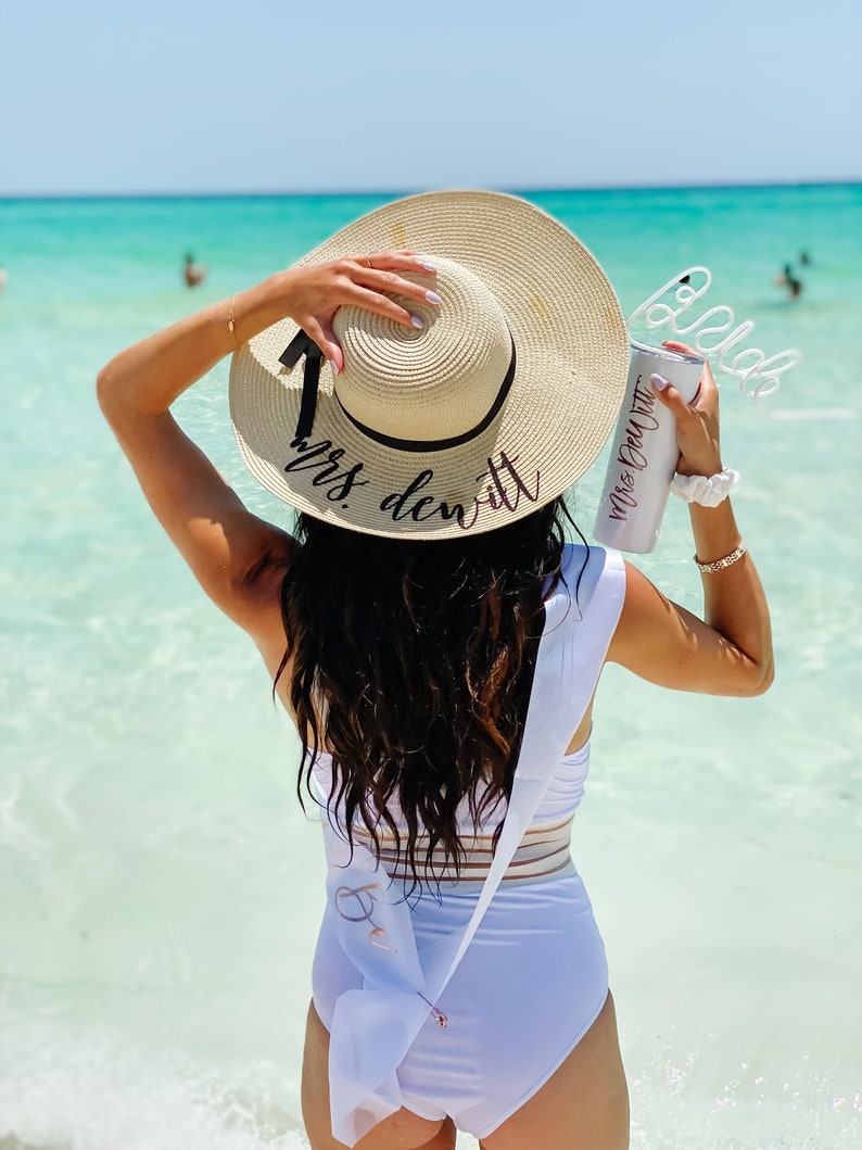 May include: A woman wearing a white swimsuit with a sash that says "Bride" and a straw hat that says "Mrs. DeWitt". She is holding a white tumbler with a straw that says "Mrs. DeWitt".