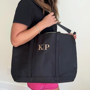 May include: A black tote bag with a gold zipper and a gold monogram of "KP" on the front. The bag has a long strap that can be worn over the shoulder.