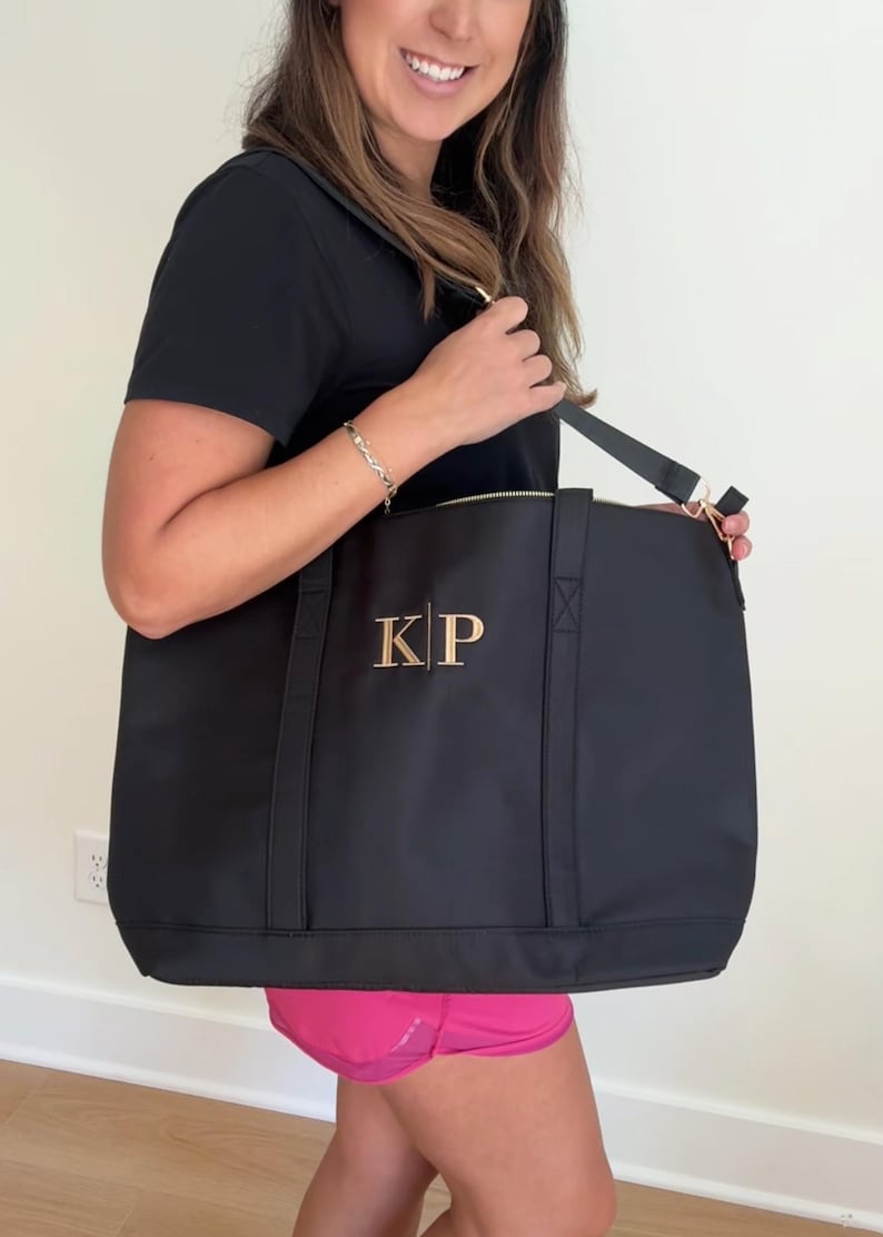 May include: A black tote bag with a gold zipper and a gold monogram of "KP" on the front. The bag has a long strap that can be worn over the shoulder.
