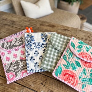 May include: Four rectangular quilted fabric pouches with fold-over flaps. The first features a pink and brown floral print, the second a blue floral pattern, the third a green and white gingham check, and the fourth a pink and green rose print.