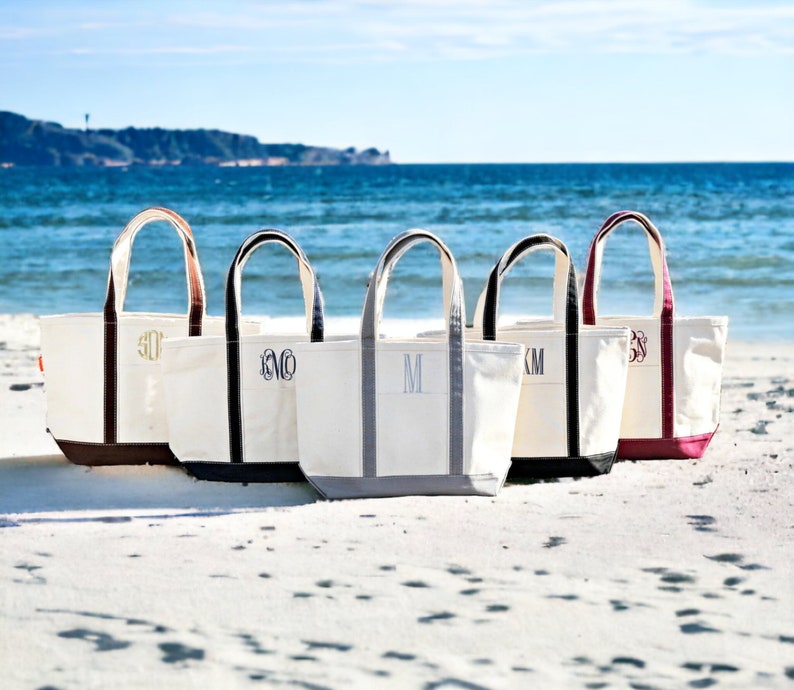 May include: Five canvas tote bags with white bodies, various colored bottoms, and handles. Each bag has a different monogram in a contrasting color. The bags are on a sandy beach with the ocean in the background.