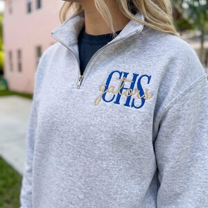Puede incluir: Una sudadera gris claro jaspeado con cremallera de un cuarto de largo en metal plateado. El pecho izquierdo muestra un bordado con las letras azules CHS y la palabra gators en cursiva dorada superpuesta sobre las letras.