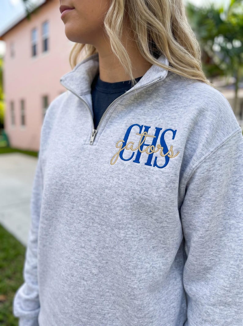 Puede incluir: Una sudadera gris claro jaspeado con cremallera de un cuarto de largo en metal plateado. El pecho izquierdo muestra un bordado con las letras azules CHS y la palabra gators en cursiva dorada superpuesta sobre las letras.