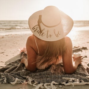 May include: A woman wearing a white sun hat with the text "bride to be" on it, sitting on a blanket on a sandy beach.
