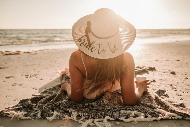 May include: A woman wearing a white sun hat with the text "bride to be" on it, sitting on a blanket on a sandy beach.