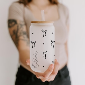 K&ouml;nnte beinhalten: Ein gefrosteter Glasbecher in Dosenform mit Holzdeckel und Strohhalm. Der Becher ist mit schwarzen Schleifen- und Herzmotiven verziert, und der Name "Olivia" ist in Schreibschrift geschrieben. Der Becher wird von einer Person gehalten.