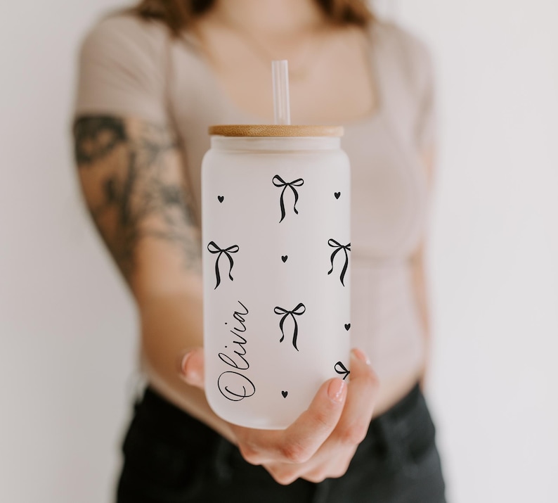 K&ouml;nnte beinhalten: Ein gefrosteter Glasbecher in Dosenform mit Holzdeckel und Strohhalm. Der Becher ist mit schwarzen Schleifen- und Herzmotiven verziert, und der Name "Olivia" ist in Schreibschrift geschrieben. Der Becher wird von einer Person gehalten.