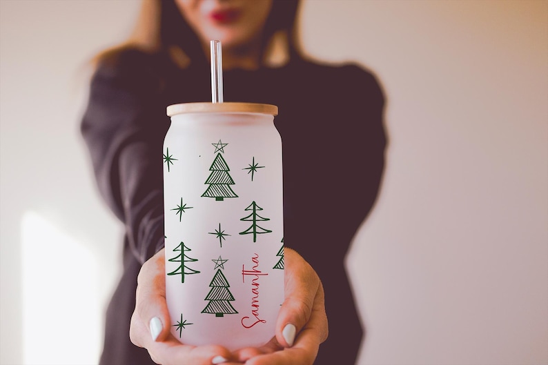 K&ouml;nnte beinhalten: Vereister Glasbecher in Dosenform mit Bambusdeckel und Strohhalm. Der Becher ist mit gr&uuml;nen Weihnachtsb&auml;umen und Sternen verziert. Der Name "Samantha" ist in roter Schrift auf der Seite geschrieben.