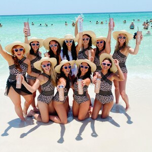 May include: A group of women wearing leopard print swimsuits and wide-brimmed hats pose on a white sandy beach. They are holding personalised tumblers with the names "Bride", "Maid of Honour", and "Bridesmaid" written on them.