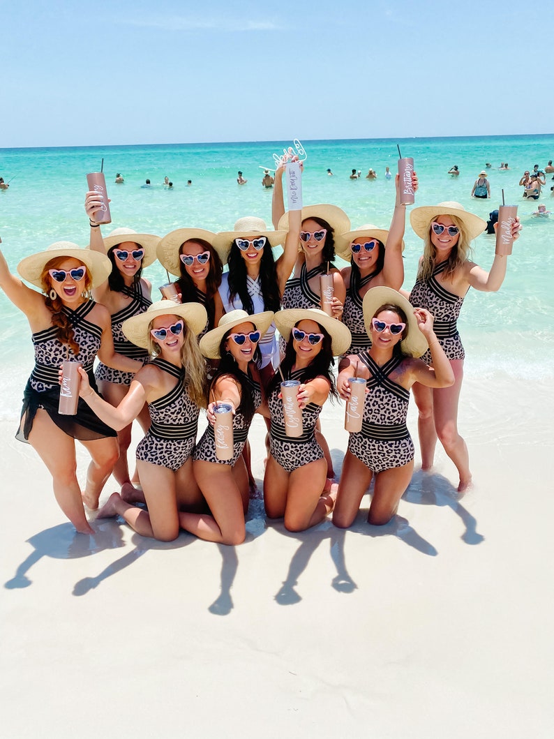 May include: A group of women wearing leopard print swimsuits and wide-brimmed hats pose on a white sandy beach. They are holding personalised tumblers with the names "Bride", "Maid of Honour", and "Bridesmaid" written on them.