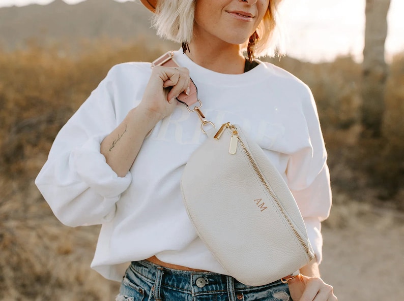 May include: A white leather fanny pack with a gold zipper and a gold chain. The fanny pack has the initials "AM" embossed on the front in a rose gold color. The fanny pack is being worn by a person with a white sweatshirt and blue jeans.