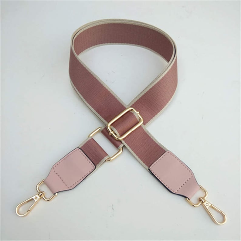 K&ouml;nnte beinhalten: Ein verstellbarer Schultergurt in Rosa und Braun mit goldenen Beschl&auml;gen und zwei drehbaren Haken. Der Gurt ist aus gewebtem Stoff mit einem wei&szlig;en Streifen.