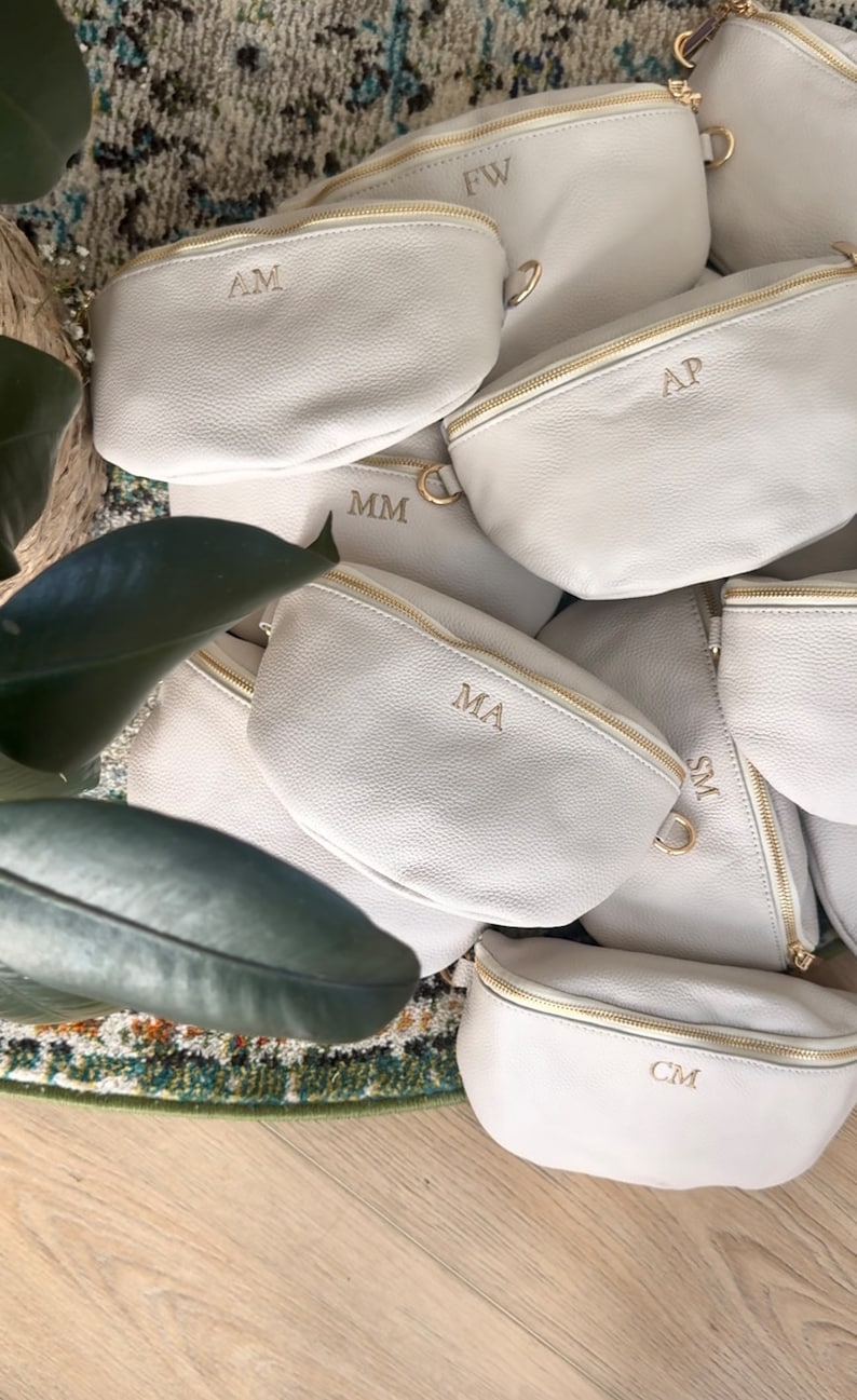 K&ouml;nnte beinhalten: Eine Gruppe von cremefarbenen Bauchtaschen mit goldenen Rei&szlig;verschl&uuml;ssen und personalisierten Initialen. Die Initialen sind AM, FW, AP, MM, MA, SM und CM.