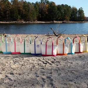 May include: A row of canvas tote bags with colorful trim and handles are displayed on a sandy beach. The bags have white bodies with colored trim in green, blue, pink, orange, yellow, and navy. Some bags have embroidered initials or text.
