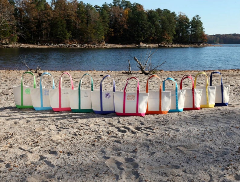 May include: A row of canvas tote bags with colorful trim and handles are displayed on a sandy beach. The bags have white bodies with colored trim in green, blue, pink, orange, yellow, and navy. Some bags have embroidered initials or text.