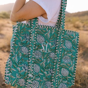 May include: A green tote bag with a floral pattern and the initials "SF" embroidered on the front. The bag has a striped border in pink and white, and the handles are also striped. The bag is made of a quilted material.