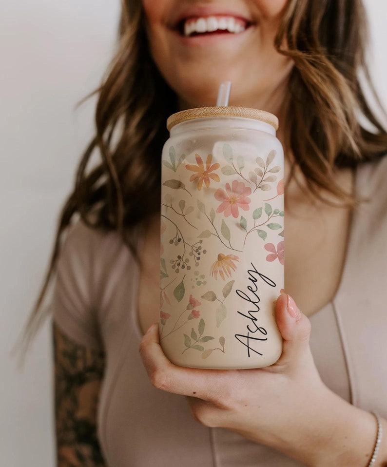 K&ouml;nnte beinhalten: Eine Glas-Getr&auml;nkedose mit floralem Design und dem Namen "Ashley" in schwarzer Schrift. Die Dose hat einen Bambusdeckel und einen Strohhalm. Das florale Design umfasst rosa, orange und gelbe Blumen mit gr&uuml;nen Bl&auml;ttern.