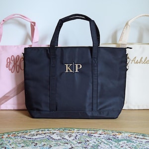 May include: Three tote bags with personalized embroidery. The bag on the left is pink with the monogram "B&H". The middle bag is black with the monogram "KP". The bag on the right is cream with the name "Ashley" embroidered on it.