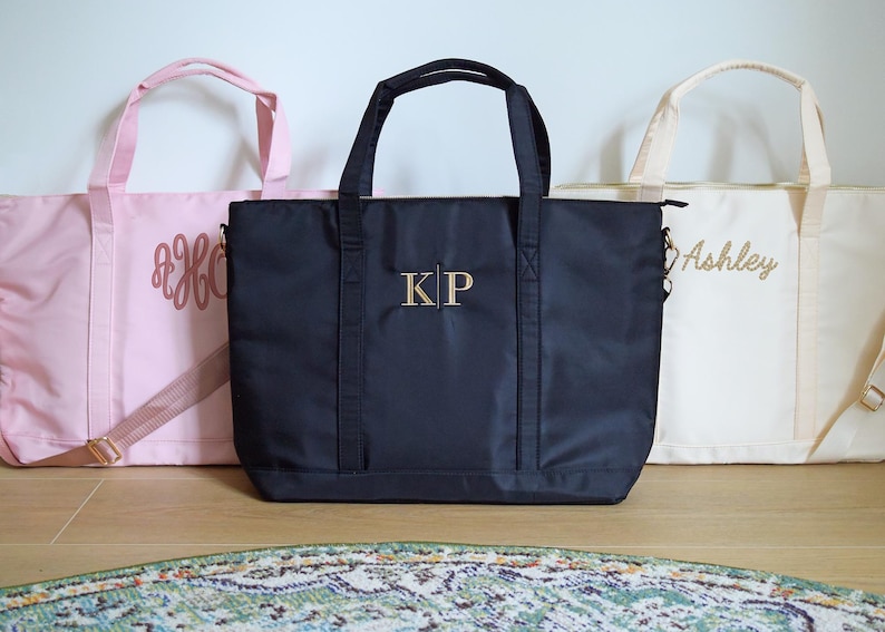 May include: Three tote bags with personalized embroidery. The bag on the left is pink with the monogram "B&H". The middle bag is black with the monogram "KP". The bag on the right is cream with the name "Ashley" embroidered on it.