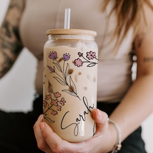 K&ouml;nnte beinhalten: Ein klares Glas mit Holzdeckel und Strohhalm, gef&uuml;llt mit einem cremigen Getr&auml;nk. Das Glas ist mit rosa und lila Blumenillustrationen und dem Namen "Sarah" in Schreibschrift verziert. Ein stilvoller und personalisierter Trinkartikel.