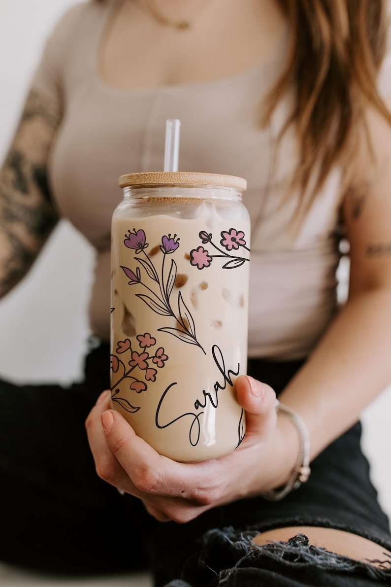 K&ouml;nnte beinhalten: Ein klares Glas mit Holzdeckel und Strohhalm, gef&uuml;llt mit einem cremigen Getr&auml;nk. Das Glas ist mit rosa und lila Blumenillustrationen und dem Namen "Sarah" in Schreibschrift verziert. Ein stilvoller und personalisierter Trinkartikel.