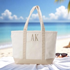 May include: A cream-colored canvas tote bag with beige accents, featuring the initials "AK" in a matching beige color. The bag has sturdy handles and a rectangular shape, perfect for carrying beach essentials. The bag is sitting on a white towel on a sandy beach.