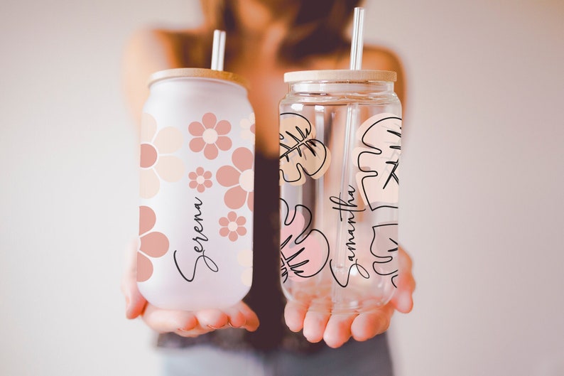 K&ouml;nnte beinhalten: Zwei Glas-Getr&auml;nkedosen mit Bambusdeckeln und Strohhalmen. Eine ist mattiert mit rosa Blumen und dem Namen "Serena". Die andere ist klar mit rosa Blattmustern und dem Namen "Samantha".