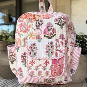 May include: A quilted backpack featuring a patchwork design with pink, orange, and green floral patterns. The bag includes a top handle, two side pockets, and a front pocket with a flap. The letters SK are embroidered in pink on the front.