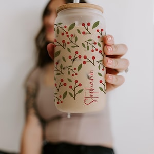 K&ouml;nnte beinhalten: Ein durchsichtiges Glas in Dosenform mit einem Holzdeckel und Strohhalm. Der Becher ist mit einem Muster aus gr&uuml;nen Bl&auml;ttern und roten Beeren verziert. Der Name "Stephanie" ist in roter Schrift auf der Seite geschrieben.