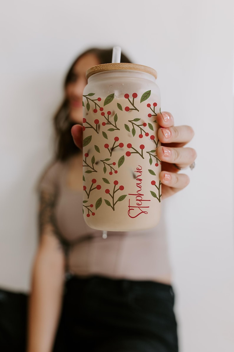 K&ouml;nnte beinhalten: Ein durchsichtiges Glas in Dosenform mit einem Holzdeckel und Strohhalm. Der Becher ist mit einem Muster aus gr&uuml;nen Bl&auml;ttern und roten Beeren verziert. Der Name "Stephanie" ist in roter Schrift auf der Seite geschrieben.