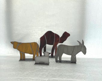 May include: A set of four animal figures crafted from stained glass with metal solder lines. The collection includes a yellow cow, a dark brown camel, a small white lamb, and a grey donkey, each mounted on a rectangular metal base.