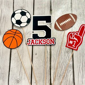 以下が含まれることがあります： スポーツをテーマにしたケーキトッパー5個。白黒のサッカーボール、オレンジ色のバスケットボール、赤い文字でジャクソンと書いてある黒い数字の5、茶色と白のアメリカンフットボール、そして赤いフォームフィンガーに1の数字。