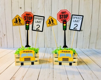 May include: Two yellow school bus centerpieces with the name Preston on the front. Each centerpiece has a wooden crate with green grass and a variety of traffic signs including a stop sign, a speed limit sign, and a school crossing sign.