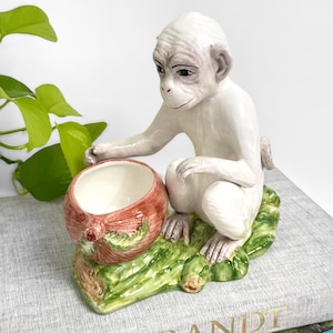 May include: A ceramic monkey figurine with a white body and a red and brown bowl. The monkey is perched on a green base. The figurine is sitting on a book with the word "REMBRANDT" on the cover.