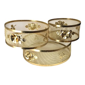 May include: Three gold-colored, mesh-style decorative baskets. Each basket is round with a floral design on the side. The baskets are stacked, with the largest at the bottom and the smallest at the top.