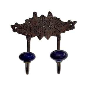 May include: A decorative wall hook with two dark blue ceramic knobs and two hooks. The top portion is a dark brown, ornate metal design with a leaf and vine pattern. The hooks are made of dark metal.