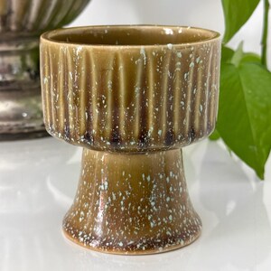 May include: A pedestal planter with a brown glaze and vertical ribbed texture on the bowl. The planter has a speckled pattern of white and light blue. The base is a flared shape.