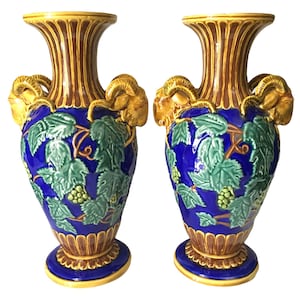 May include: A pair of decorative ceramic vases. The vases feature a blue body with green grape leaf and cluster designs. Gold ram's head handles and a yellow rim and base complete the design. The vases are approximately 12 inches tall.
