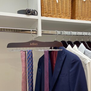 May include: A wooden tie rack with the name "Jeffrey" engraved on it. The rack has 10 metal hooks for hanging ties. There are three ties hanging on the rack, one pink, one red with white polka dots, and one blue with white squares.