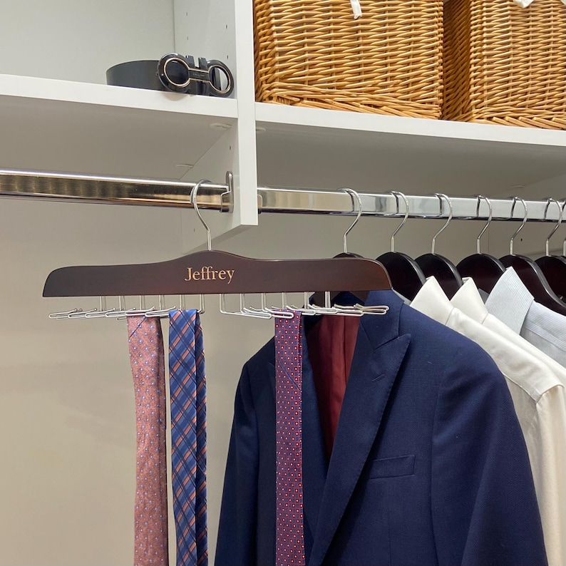 May include: A wooden tie rack with the name "Jeffrey" engraved on it. The rack has 10 metal hooks for hanging ties. There are three ties hanging on the rack, one pink, one red with white polka dots, and one blue with white squares.