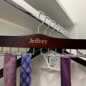 May include: A dark brown wooden tie rack with the name "Jeffrey" engraved on it. The rack is holding several patterned ties, including a blue and white plaid tie, a red and blue patterned tie, and a pink and blue patterned tie.