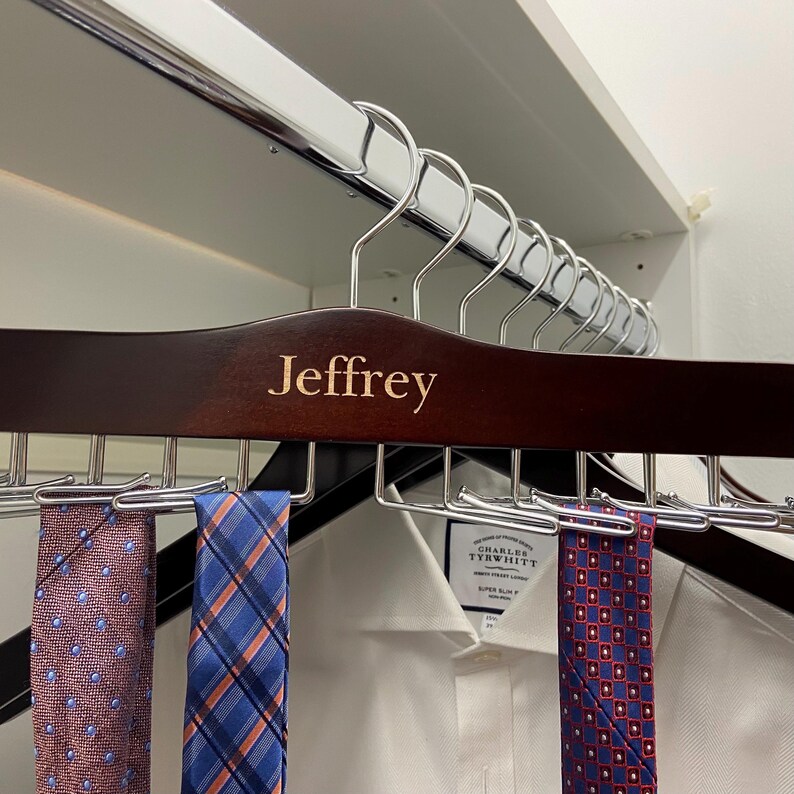 May include: A dark brown wooden tie rack with the name "Jeffrey" engraved on it. The rack is holding several patterned ties, including a blue and white plaid tie, a red and blue patterned tie, and a pink and blue patterned tie.