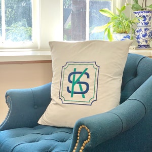 May include: A white cushion with a navy blue and green monogram "KS" in a decorative frame. The cushion is sitting on a dark blue armchair with gold-coloured nailhead trim.
