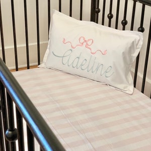 May include: A white pillow with a pink bow and the name "Adeline" embroidered in blue thread. The pillow is on a white and pink striped cot sheet.