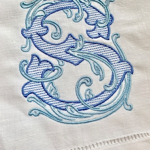 May include: A blue and white embroidered letter S with a decorative floral design. The letter is stitched on a white linen fabric.
