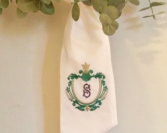 May include: White fabric with a green and gold embroidered crest featuring a purple letter "S" in the center.